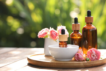 Fototapeta na wymiar Bottles of rose essential oil and flowers on wooden table outdoors, space for text