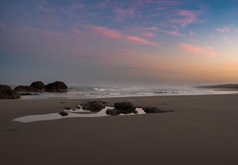 Sunrise over the Oregon Coastline, with waves washing over rough rockers to reach the sandy beach.