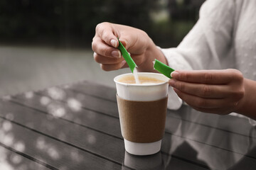 Woman adding sugar to aromatic coffee in paper cup at table, closeup