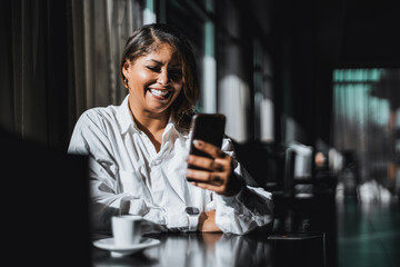 A portrait of a laughing beautiful plus-size Hispanic woman entrepreneur laughing while having a video call with a colleague via smartphone while sitting in a dark cafe interior during a coffee break