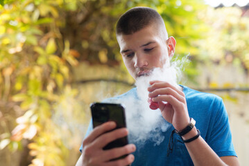Young man inhaling a smoke of a vape, or electronic cigarette, autumn surrounding - obrazy, fototapety, plakaty