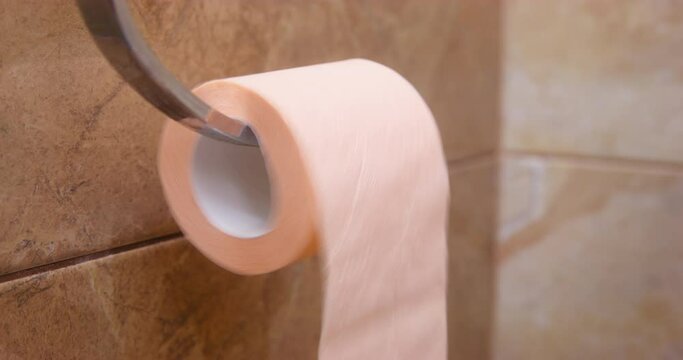 Toilet paper unrolled fast on metal stick and becomes smaller against beige tiled wall. Person manages to stop roll of paper by finger closeup