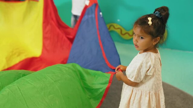 Little dark-skinned girl in dress holding one corner of rainbow play parachute during playtime in nusery. Multiracial group. Horizontal indoor shot. High quality 4k footage