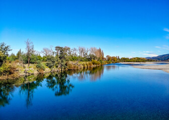 A landscape image of trees reflecting in the still blue waters of the Wairau River on the Marlborough Region of the South Island of New Zealand