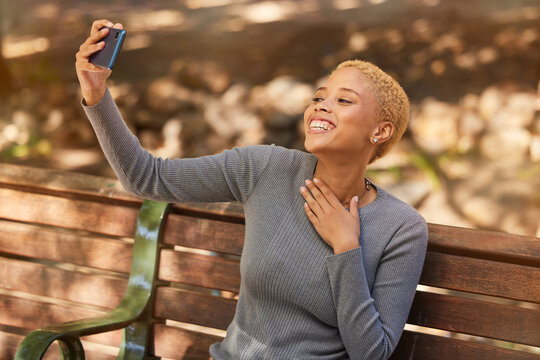 Park bench, phone selfie and black woman in nature outdoors taking picture. Happy, smile and girl from South Africa with 5g mobile for photo, social media post or comic memory alone at park outside.