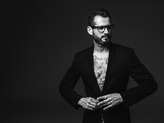 Portrait of handsome confident stylish hipster lambersexual model. Sexy modern man dressed in black jacket. Naked torso with tattoos.Fashion male posing in studio on dark background. In spectacles