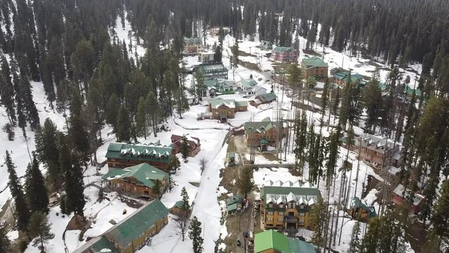 Aerial slow movement around snow covered forest with buildings and dense pine trees in Gulmarg