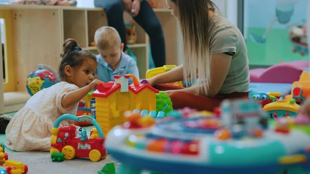 Multiracial group of little children playing with various colorful toys with their daycare teacher. Nursery playtime. Horizontal indoor shot. High quality 4k footage