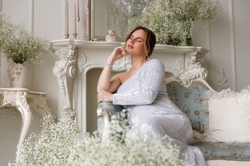 beautiful pregnant woman in evening dress sits on a sofa in a white room. The concept of motherhood...