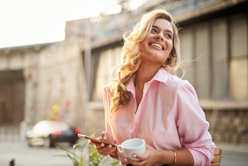 Happy young woman holding a cup of coffee and looking at her cell phone, typing a message or...