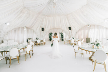 the bride stands with her back in the middle of a large white tent for a wedding banquet in white.