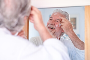 Obraz premium Skin care. Handsome senior man applying cream at his face and looking at himself with smile while standing in front of the mirror