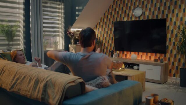 Couple on sofa in living room, watching action movie on TV or criminal blockbuster on streaming service, talking and discussing acting, resting at home on weekend. Home cinema in modern apartment.