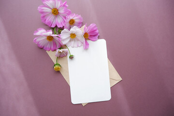 Beautiful autumnal flowers composition. Pink and white cosmos flowers with blank greeting card on pink brown background. Flat lay, top view, copy space. Autumn Greeting concept floral background.