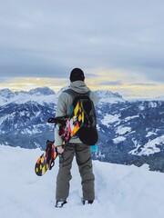 Naklejka premium Vertical shot of a snowboarder looking at the snowy mountains with a snowboard under his arm