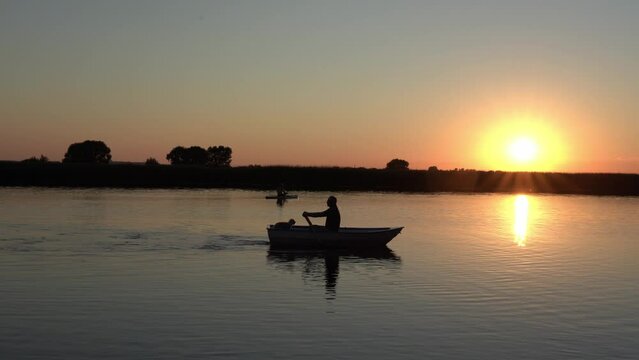An elderly man with a poodle dog is rowing a rowing boat at dusk in the late evening against the backdrop of the sunset