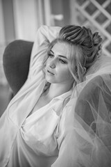 Young beautiful bride with wedding hairdo and makeup in the morning wearing a white silk negligee in the bedroom. wedding morning