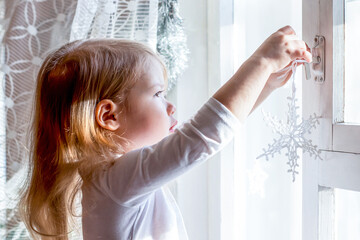 Happy little girl decorates the window with Christmas ball and snowflakes. Atmosphere of holiday, cozy, anticipation of the new year and Christmas