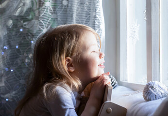 little blonde girl looking thoughtfully out the window. Cone and Christmas tree ball lying on the...