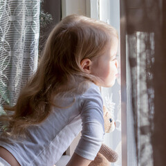 little blonde girl and her toy teddy bear looking out the window together, sticking their noses into the glass