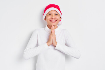 Portrait of smiling Asian man in christmas hat keeping palms together, making request isolated on white background