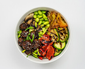 Top view of tofu poke bowl with basmati rice, edamame beans, warm zucchini and peppers in white ceramic bowl on gray background