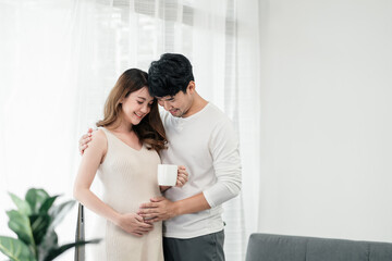 Fototapeta premium A happy asian couple. A beautiful pregnant wife is holding a cup of milk for the baby with her husband smiling and both of their hands rubbing the wife's tummy in the living room.