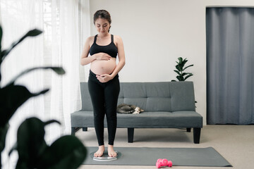 Obraz premium Pregnant Asian woman healthy standing on scales and rubbing her belly at home. Concept health care during pregnancy.