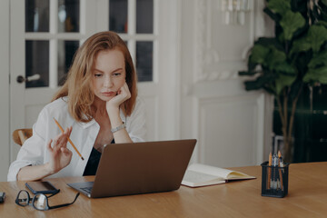 Focused female employee sitting at desk with laptop working online, thinking of problem. Remote job
