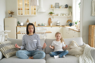 Mother with little foster daughter doing yoga exercises, sitting in lotus position on sofa together
