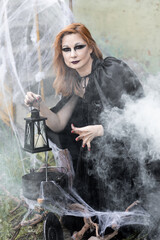 Attractive beautiful woman in a Halloween costume black cloak and scary make-up. Young girl in a look of a witch dressed up for a party making fairy potion holding black lantern