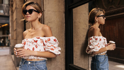 Obraz premium Calm young caucasian girl holding paper cup of coffee, looks away to street in summer. Lady blonde wears sunglasses, blouse and jeans. Rest time concept