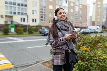 Fototapeta premium stylish young woman in an autumn coat walks around the city Listening to music in headphones with a phone in her hands