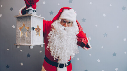 Santa Claus isholding lantern and bag of gifts looking at camera on starry background standing...