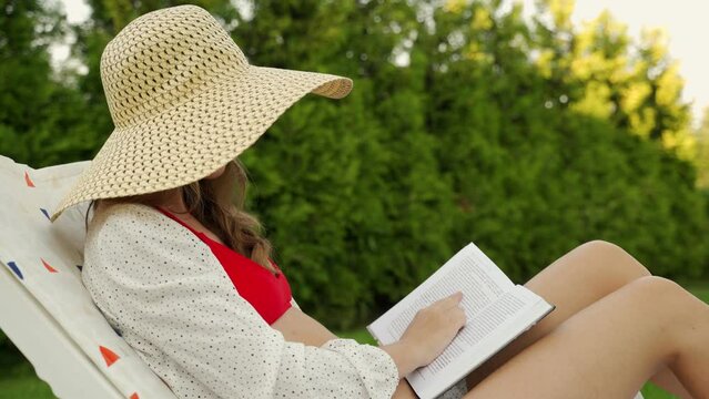 Young romantic woman in a straw hat reading a book sitting on a chaise longue in the backyard against a background of greenery. 