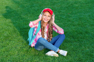 Happy schoolgirl listening to music sitting on green grass outdoors, school