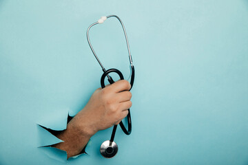 Stethoscope in a male hand on a blue background with a hole, medical concept