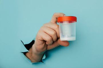 Male hand holds a plastic jar for urine tests, the hand is sticking out of a torn hole in a blue background