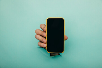 A male hand sticking out of a hole from a blue background, holds a smartphone with a black screen