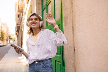 Happy young caucasian woman listening to song in headphones and using mobile phone walking. Blonde girl wears shirt with jeans. Technology concept