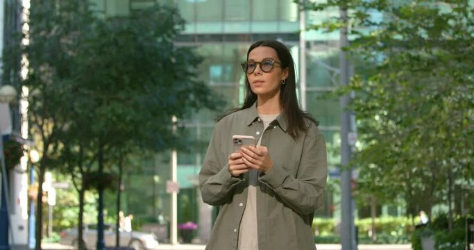 Outdoor shot of pretty woman with dark long hair holding smartphone on the street, surfing social media considerately 