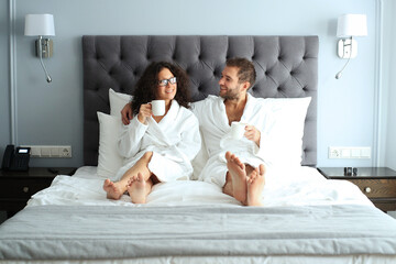 Happy couple in white bathrobe with cup of coffee on a bed in a hotel. Morning in the hotel room. - obrazy, fototapety, plakaty