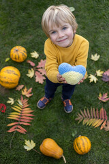 Fototapeta premium smiling boy standing among orange pumpkins and autumn leaves holds out yellow-blue Ukrainian heart