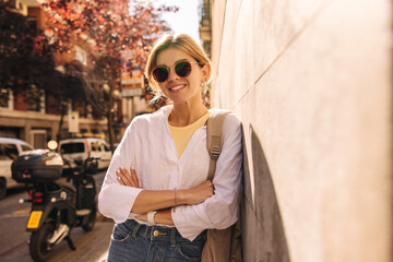 Fototapeta na wymiar Attractive young caucasian woman model in sunglasses posing leaning against city wall. Fair-haired lady wears casual light clothing in warm weather. Spring concept