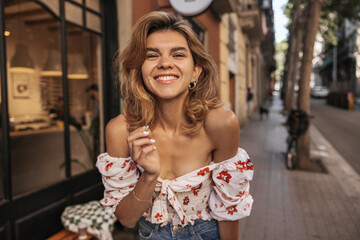 Cheerful young caucasian woman smiles teeth looking at camera walking around city during day. Blonde with wavy hair wears casual summer clothes. Happiness lifestyle concept.