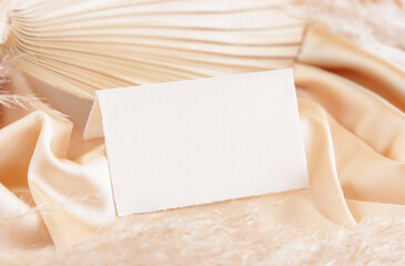 Folded place card on beige satin fabric and dried palm leaf close up, greeting or wedding mockup