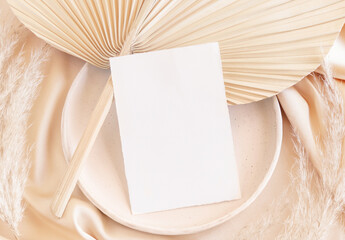 Blank card on plate on dried palm leaf and beige silky fabric close up, greeting or wedding mockup