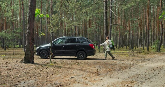 Photo from distance beautiful black car in the middle of forest woods. Exhauested tired young girl lady opening truck putting travelled backpack in sitting in car after long jorney .