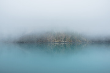Simple meditative landscape with turquoise lake in thick fog. Tranquil view through clearance in dense fog to steep slope in fading autumn colors and mirror mountain lake. Reflective azure alpine lake