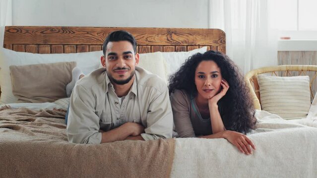 Multiracial multiethnic married couple boyfriend and girlfriend husband and wife man and woman smiling happy with real estate relocation house lie on comfortable bed in home bedroom looking at camera